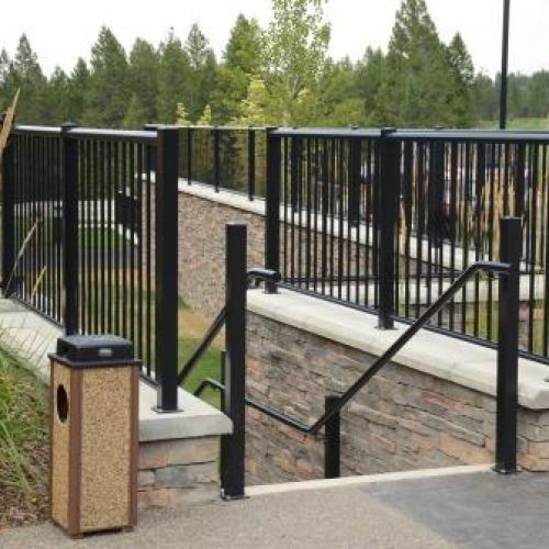 A set of DekSmart black pipe hand railings along the top of a stone deck that lead down a set of stone steps, with green trees in the background